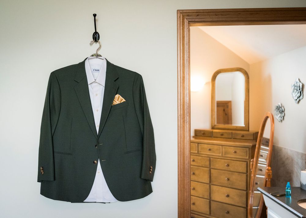 Dark green wedding suit jacket with gold pocket square hanging on a wall hook in a rustic room.