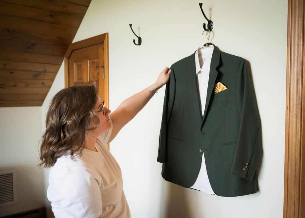 Bride in white shirt admiring her green wedding suit jacket with gold pocket square hanging on a rustic wall hook.