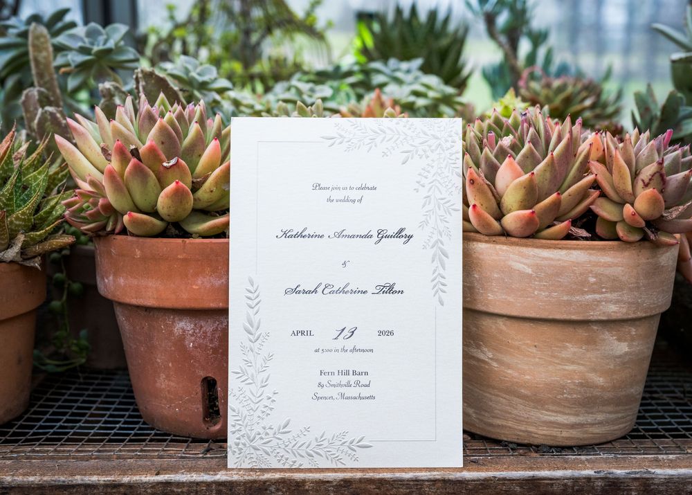 Elegant white wedding invitation with floral border for Katherine and Samuel displayed among terracotta pots of succulents.