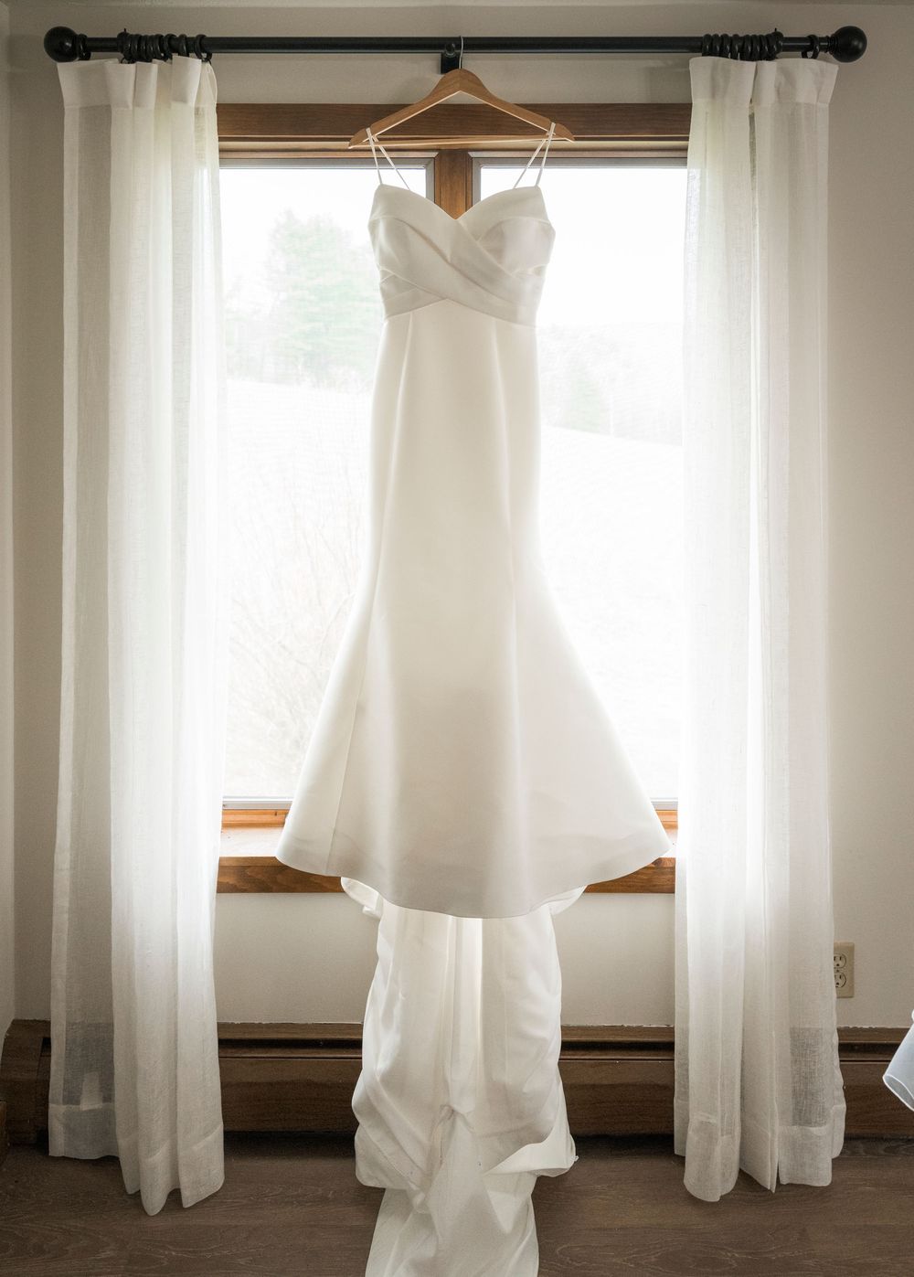 Classic white strapless mermaid wedding dress with sweetheart neckline hanging in a sunlit window with sheer curtains.