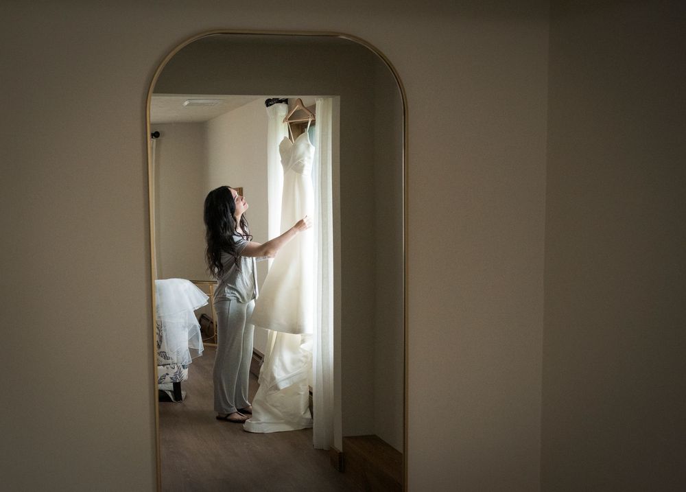 A bride views her white wedding gown hanging by a window in a softly lit bridal suite.