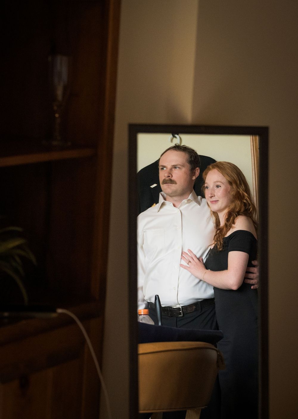 Couple sharing an intimate moment reflected in a mirror, woman in black dress embracing man in white shirt indoors.