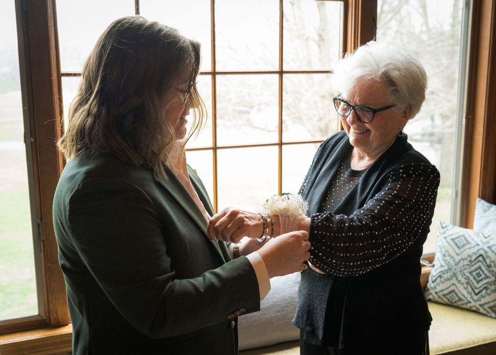 A bride in a green blazer receives a bracelet from an elderly woman near a large window with garden views.