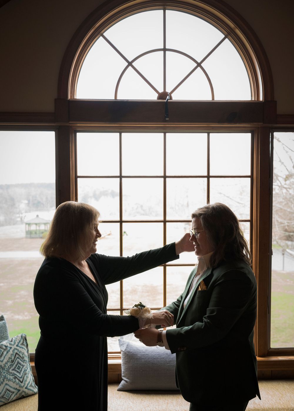 Two women sharing an emotional moment by arched window in soft natural light at wedding.
