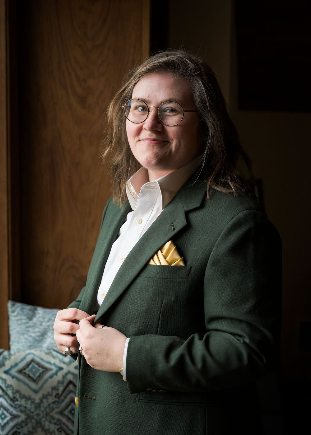 Person in green blazer with yellow pocket square and glasses posing indoors.