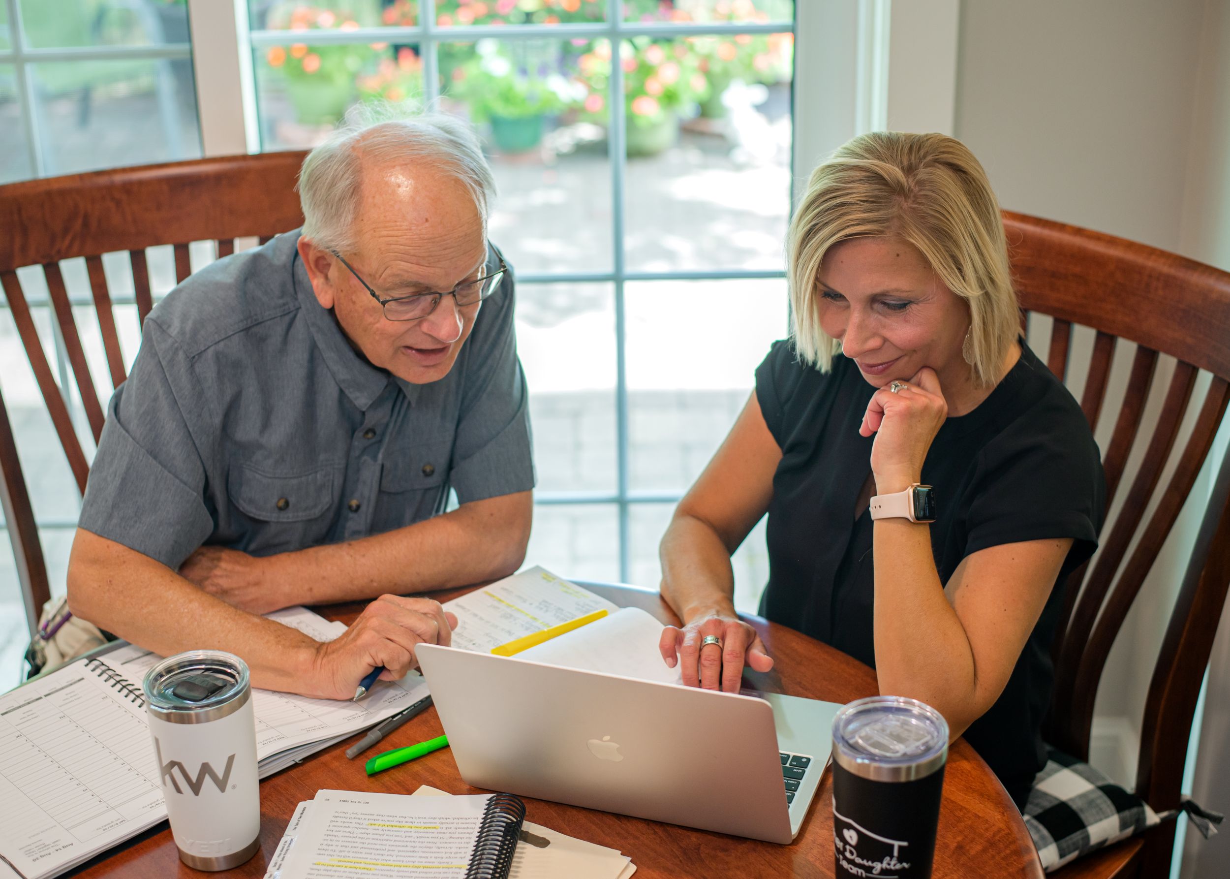 Two people reviewing work on laptop at wooden dining table.