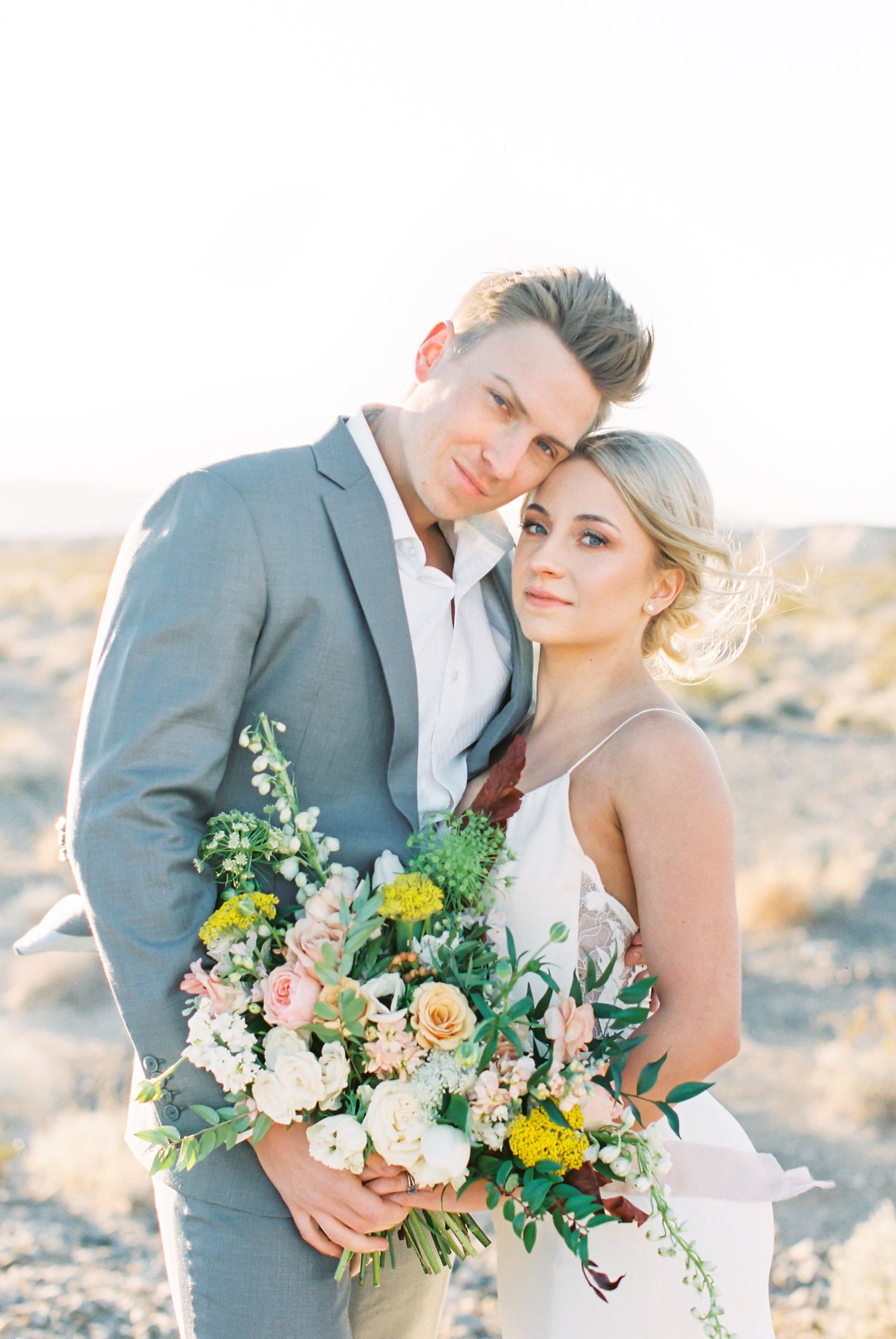 A romantic desert wedding photo session with a couple embracing and holding a delicate peach and white floral bouquet.