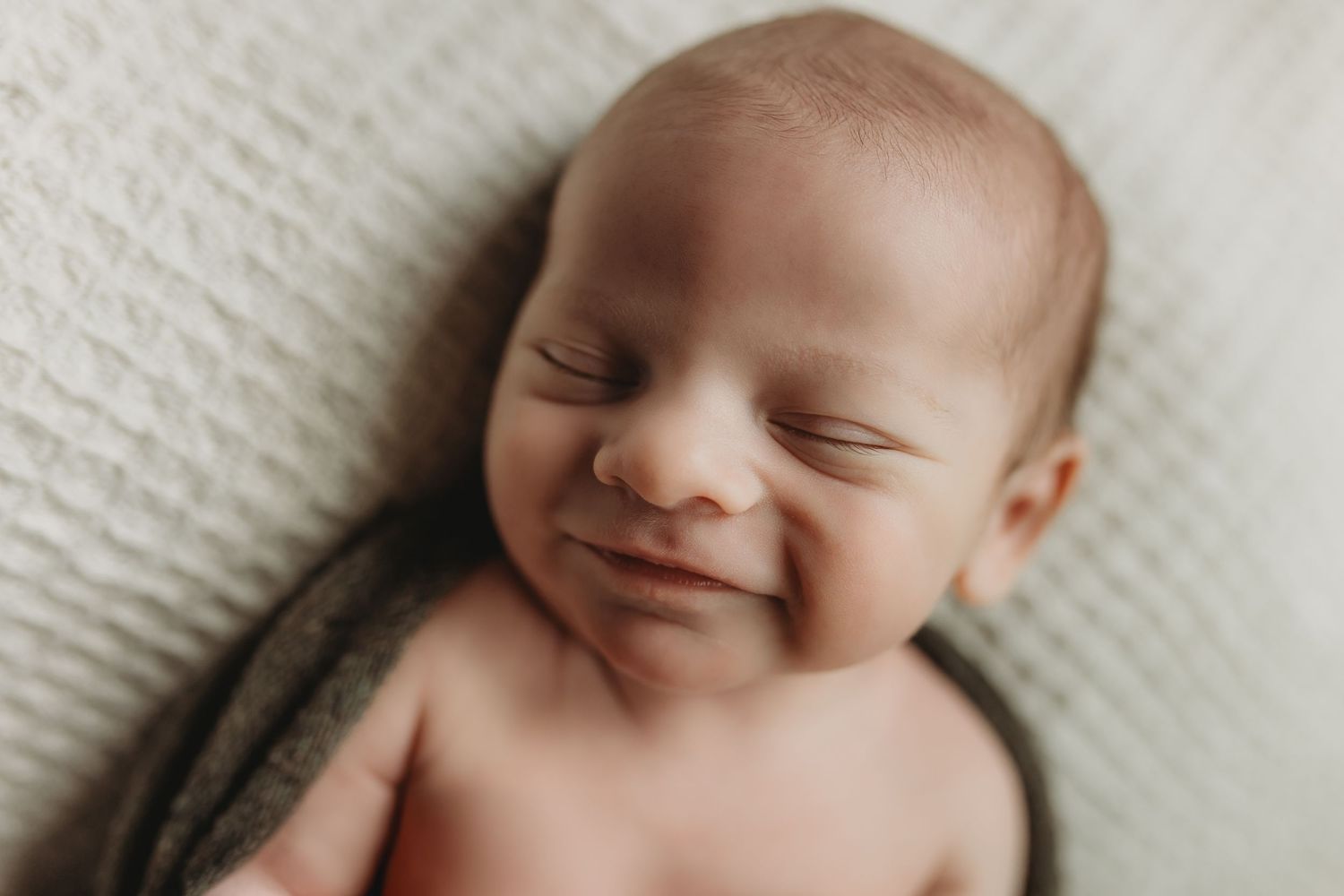 Smiling newborn baby sleeping peacefully wrapped in a soft gray blanket on a white textured background.