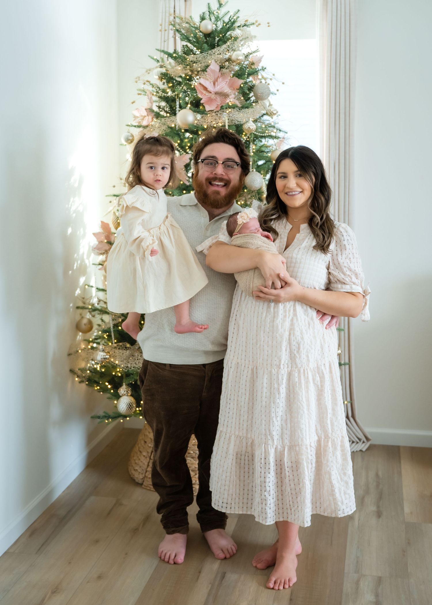 A family poses together in coordinated white and beige outfits in front of a decorated Christmas tree.