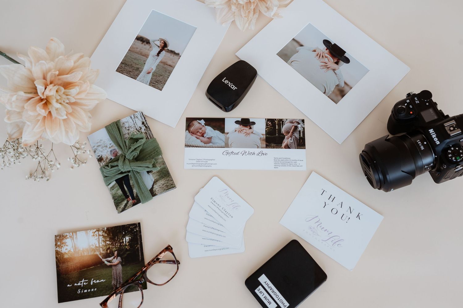 Flatlay photography setup with scattered photos, dried flowers, camera equipment and stationery on a light beige surface.