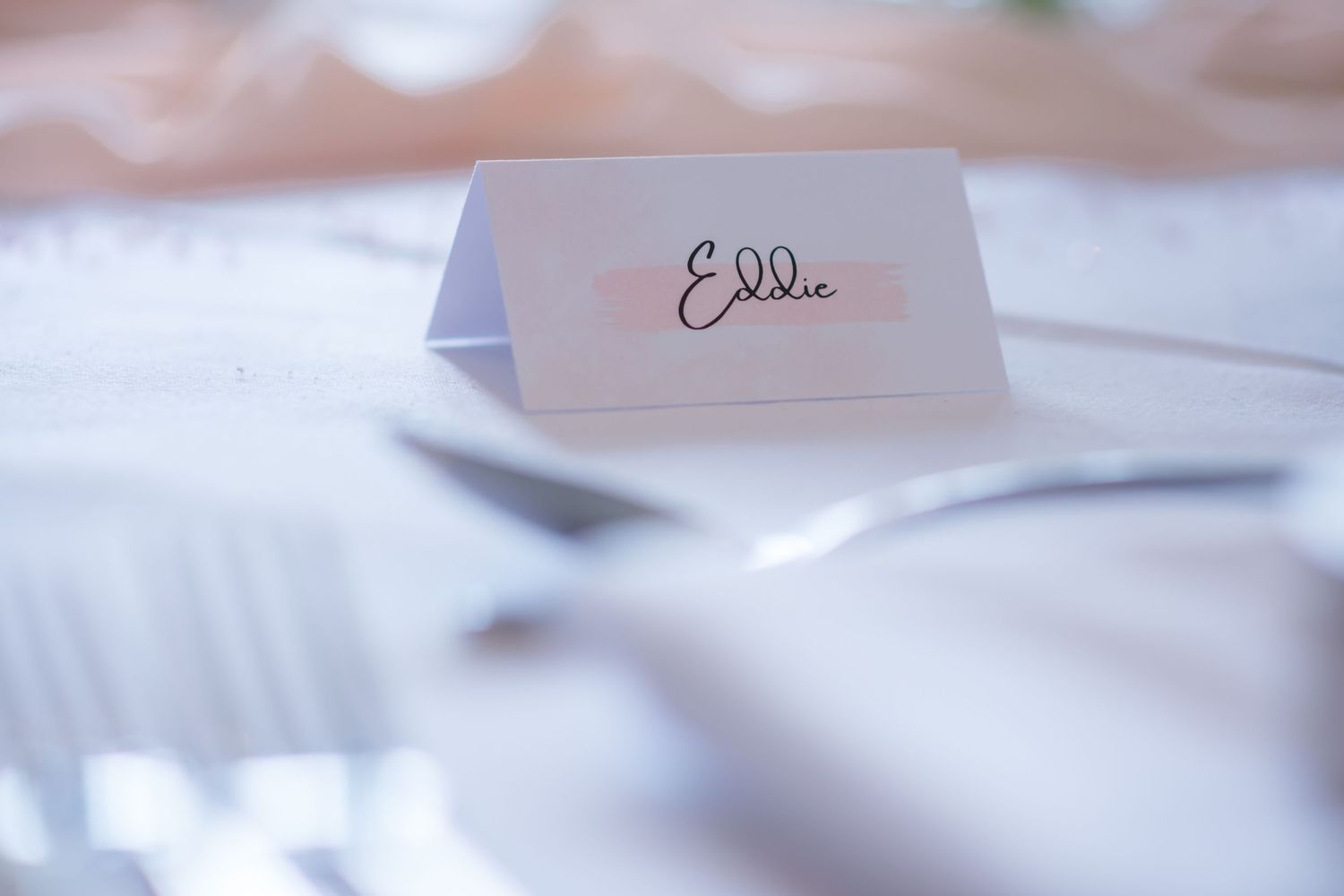 Elegant wedding place card with handwritten name sits on white linen tablecloth at formal reception setting.