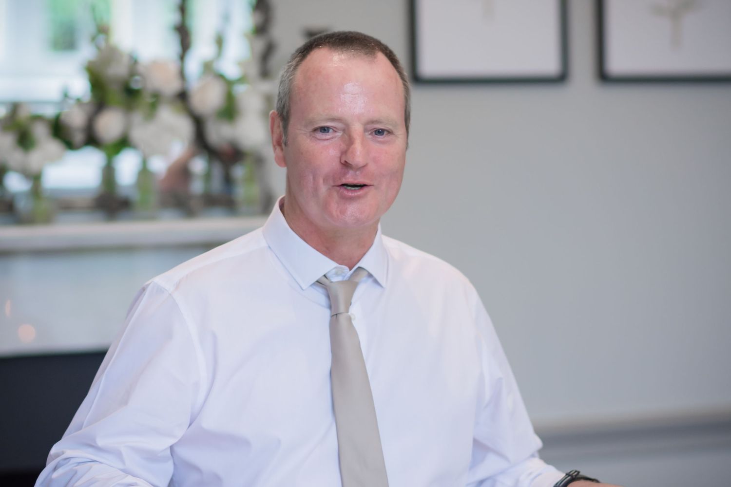Professional wearing a white dress shirt and beige tie sits at desk in modern office setting.