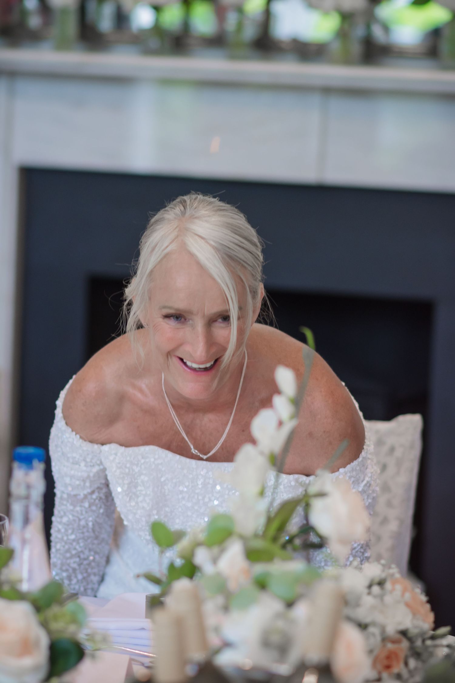Bride in a sparkling white off-shoulder gown smiles during a wedding reception with floral centerpieces.