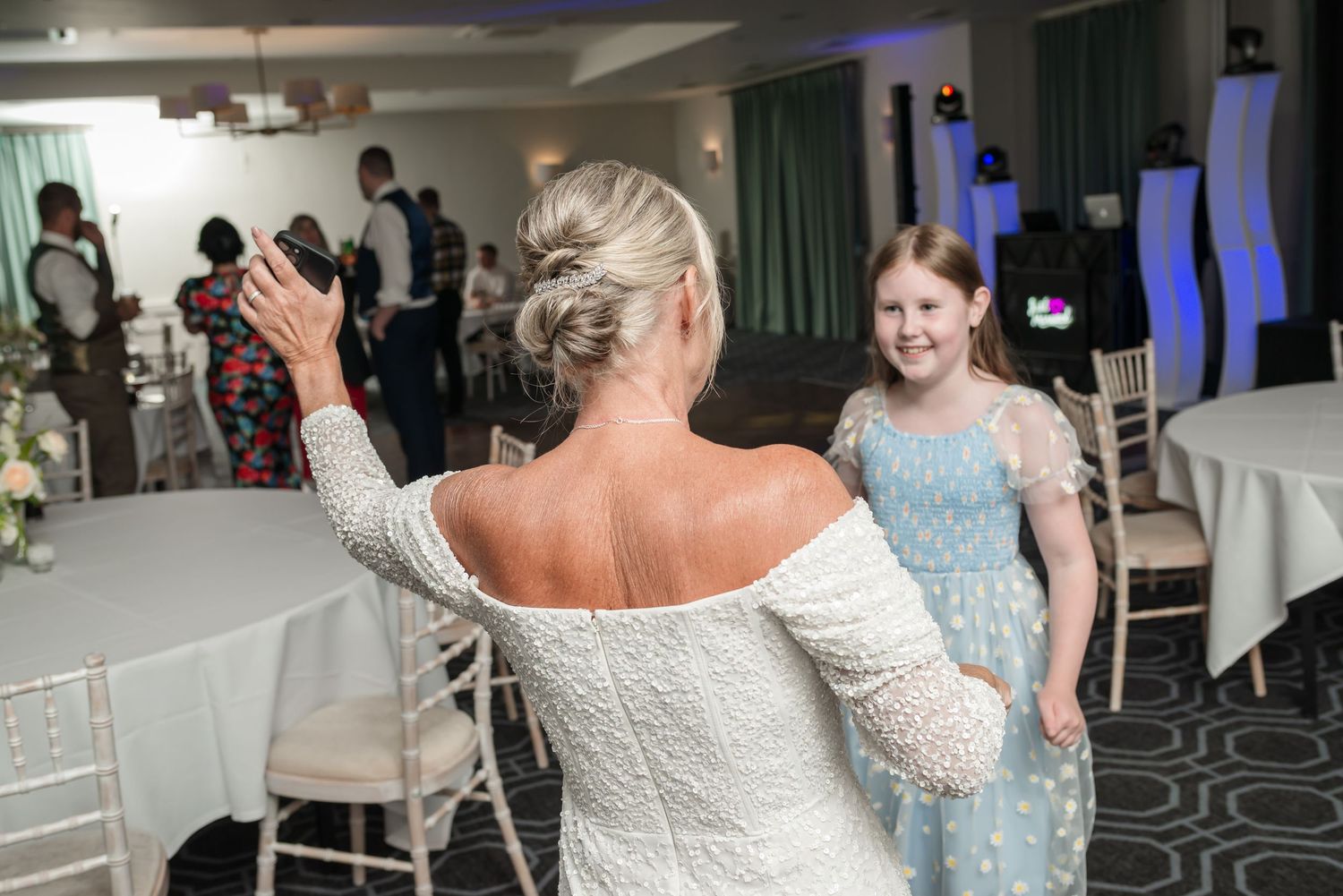 A person in a white off-shoulder dress dances at a wedding reception while a guest in a light blue dress watches.