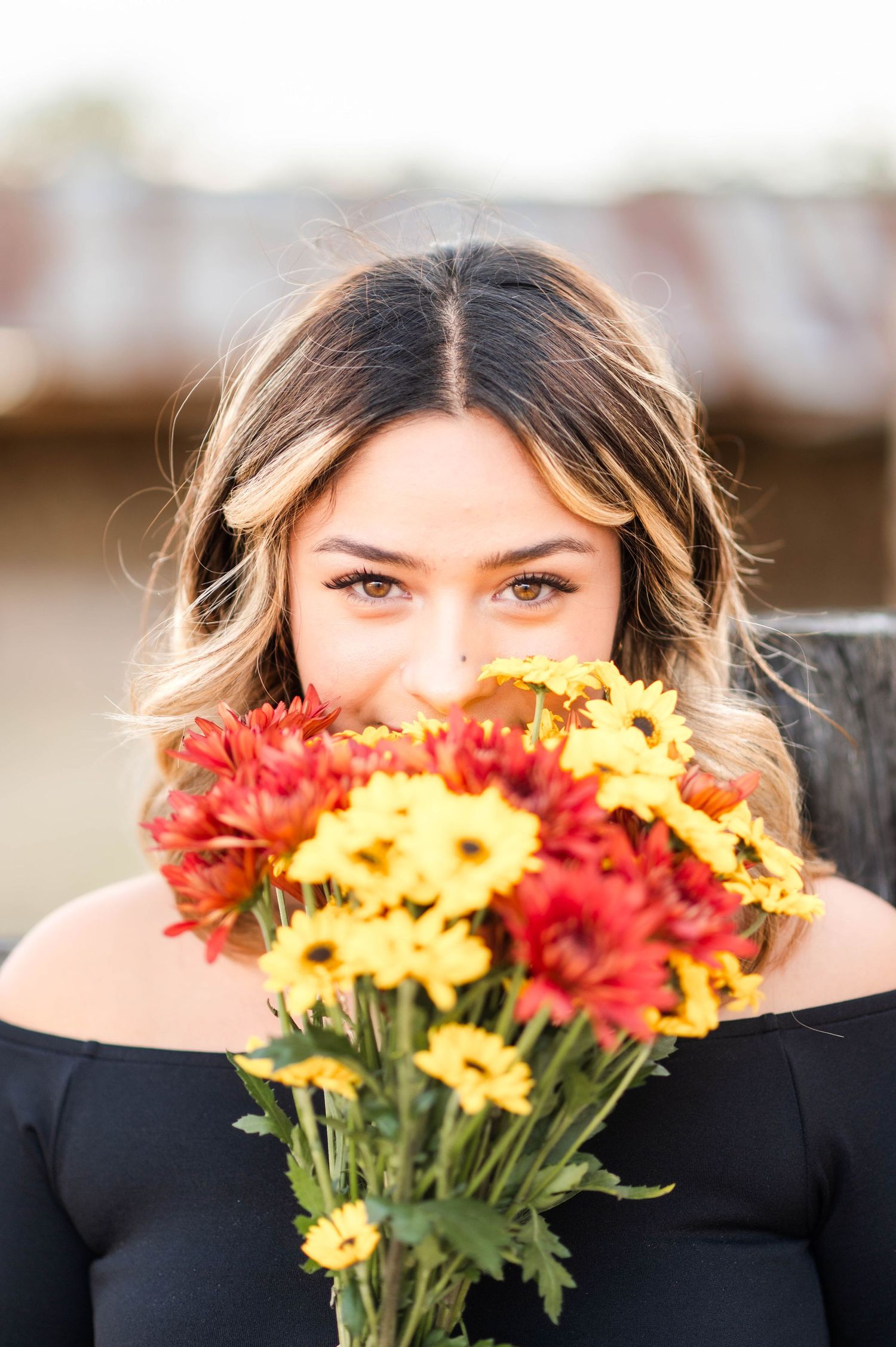 Woman holding vibrant red and yellow flowers bouquet up to her face with expressive brown eyes visible above blooms.