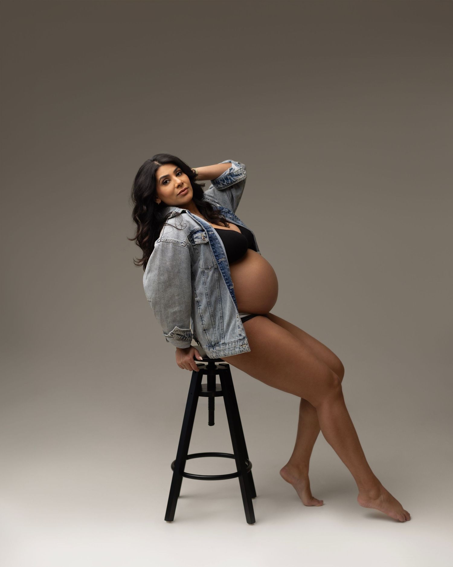 A model in a denim jacket and dark bodysuit poses on a black stool against a neutral studio backdrop.