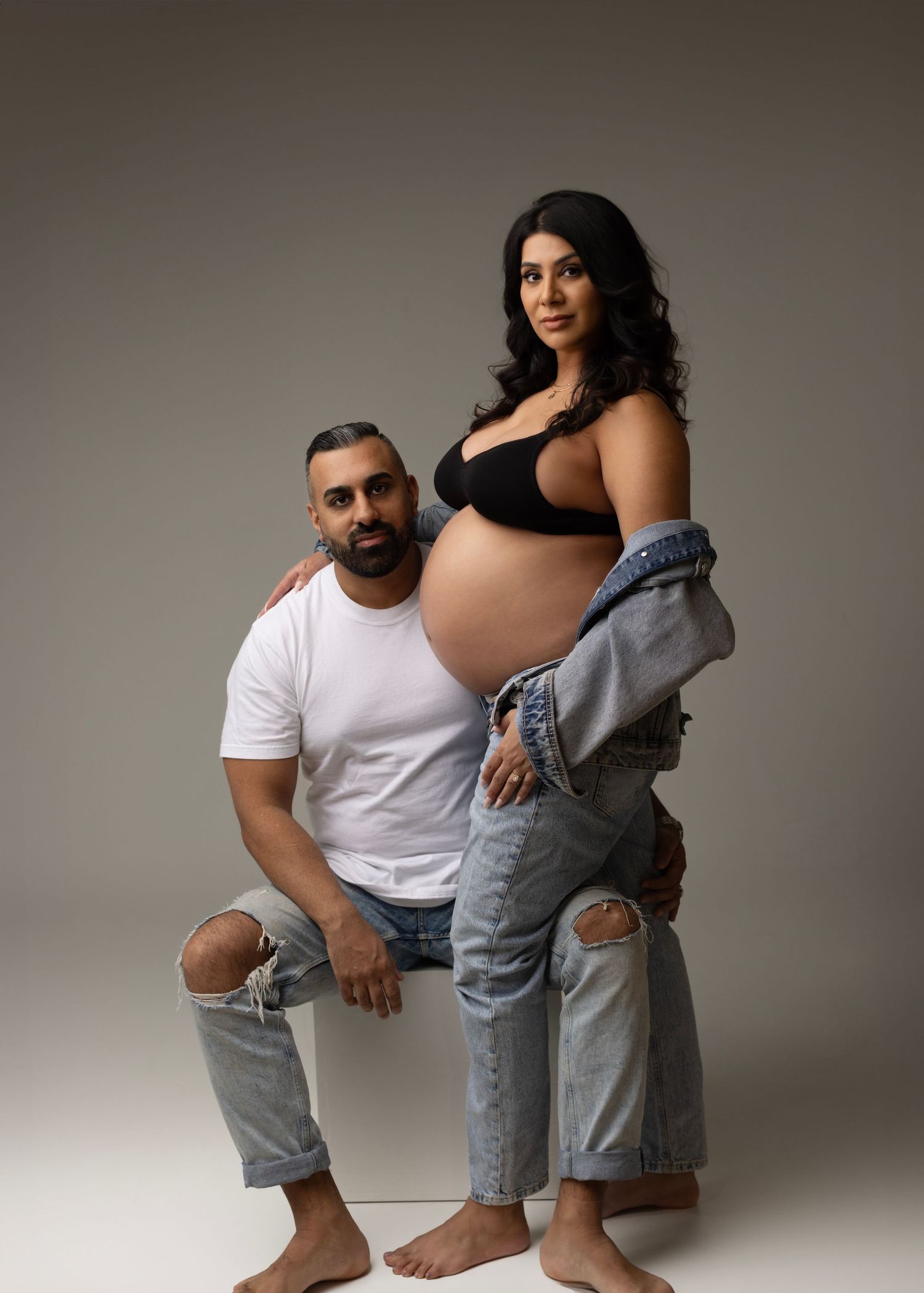 A couple poses in casual white and denim outfits during a maternity photoshoot against a neutral studio backdrop.