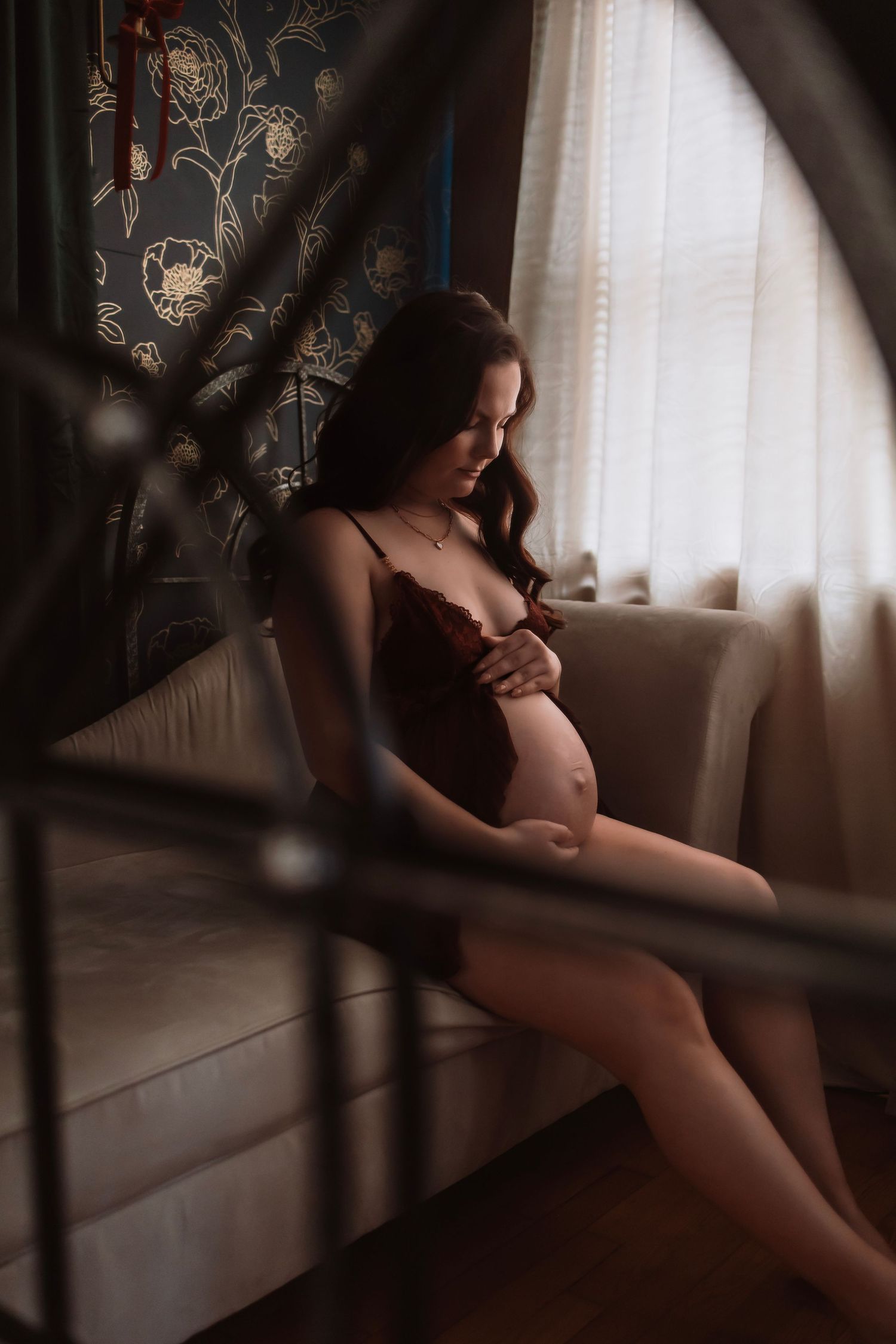 Moody maternity boudoir photo of a pregnant woman wearing lingerie sitting near a window, photographed in a warm, soft, natural light studio