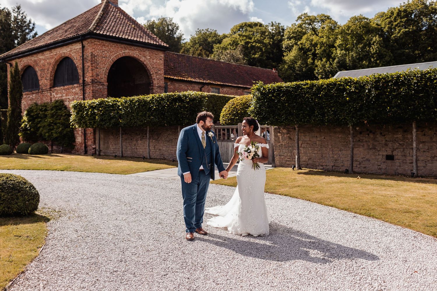 A couple walks along a white gravel path surrounded by historic brick walls and green landscaping on a sunny day.