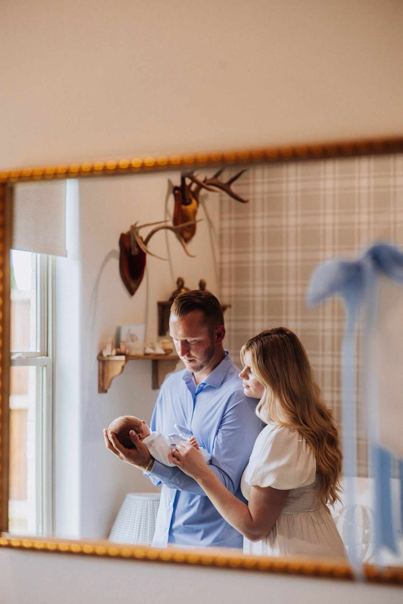 New parents admiring their newborn baby reflected in a gold-framed mirror in a cozy room.