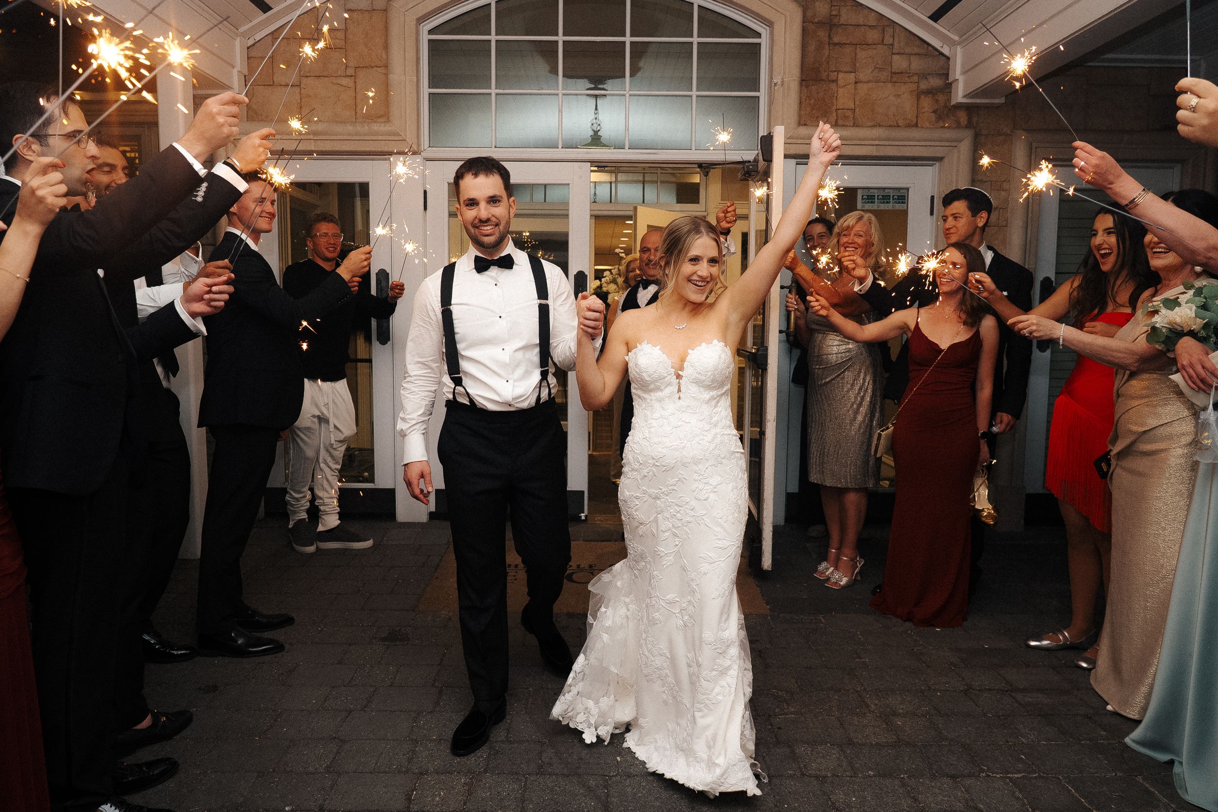 Wedding guests dance and celebrate on a reception dance floor in a white-walled venue with arched windows.