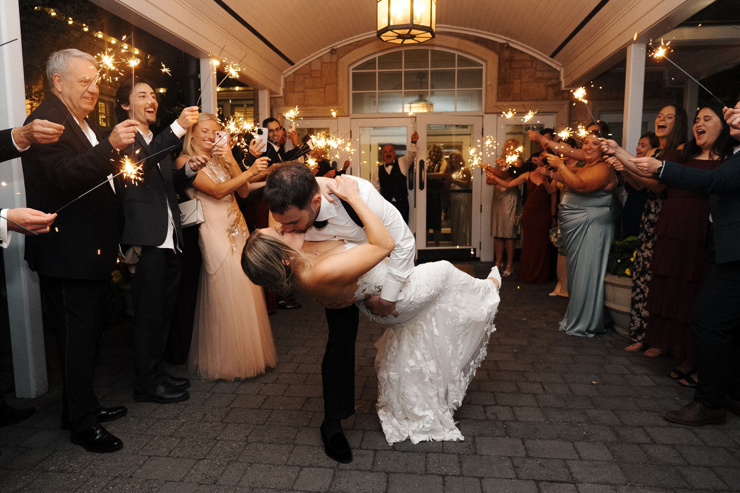 A romantic dip kiss between newlyweds surrounded by cheering guests at an evening wedding celebration.