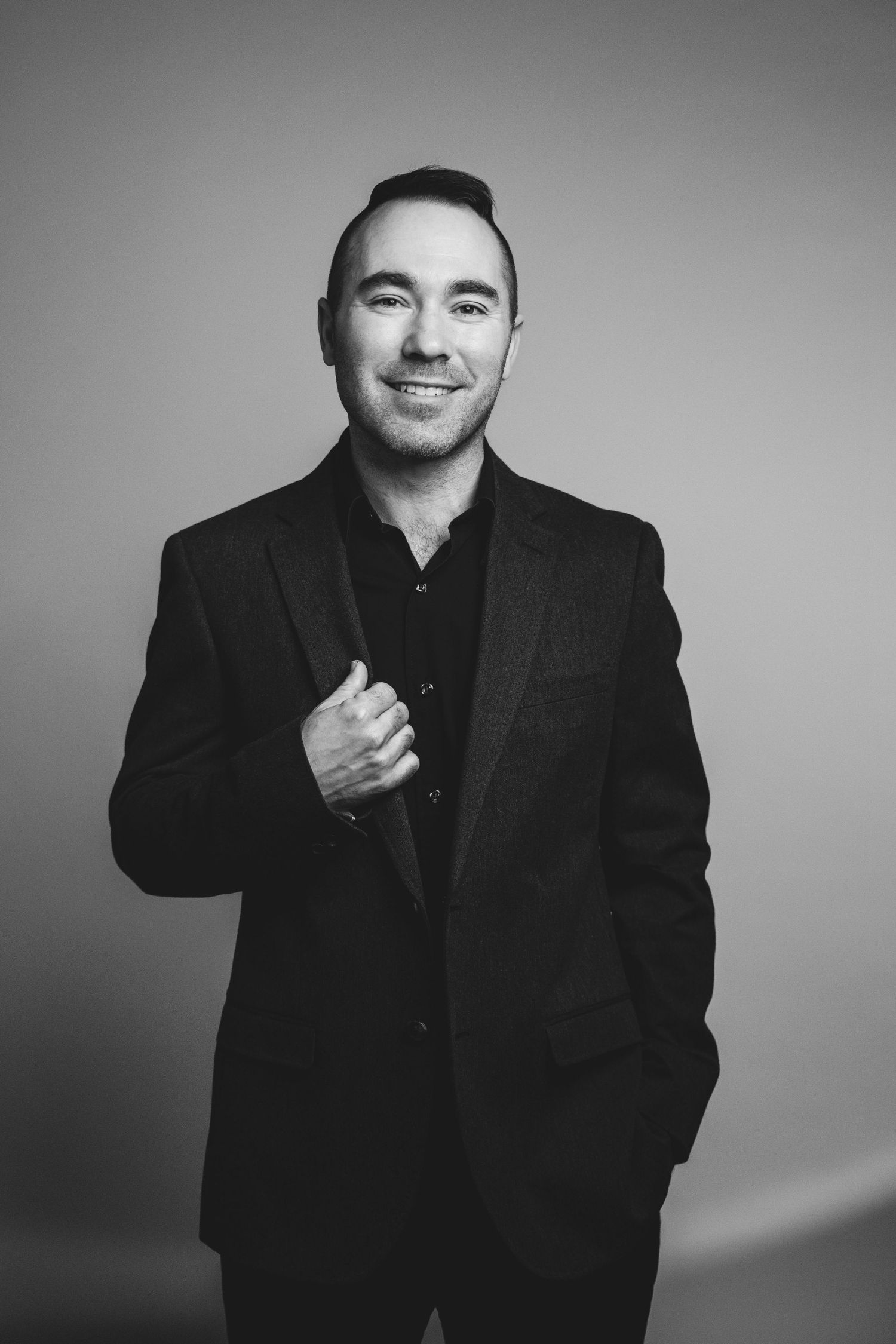 Smiling man in black suit posing confidently in a professional black and white studio portrait photo.