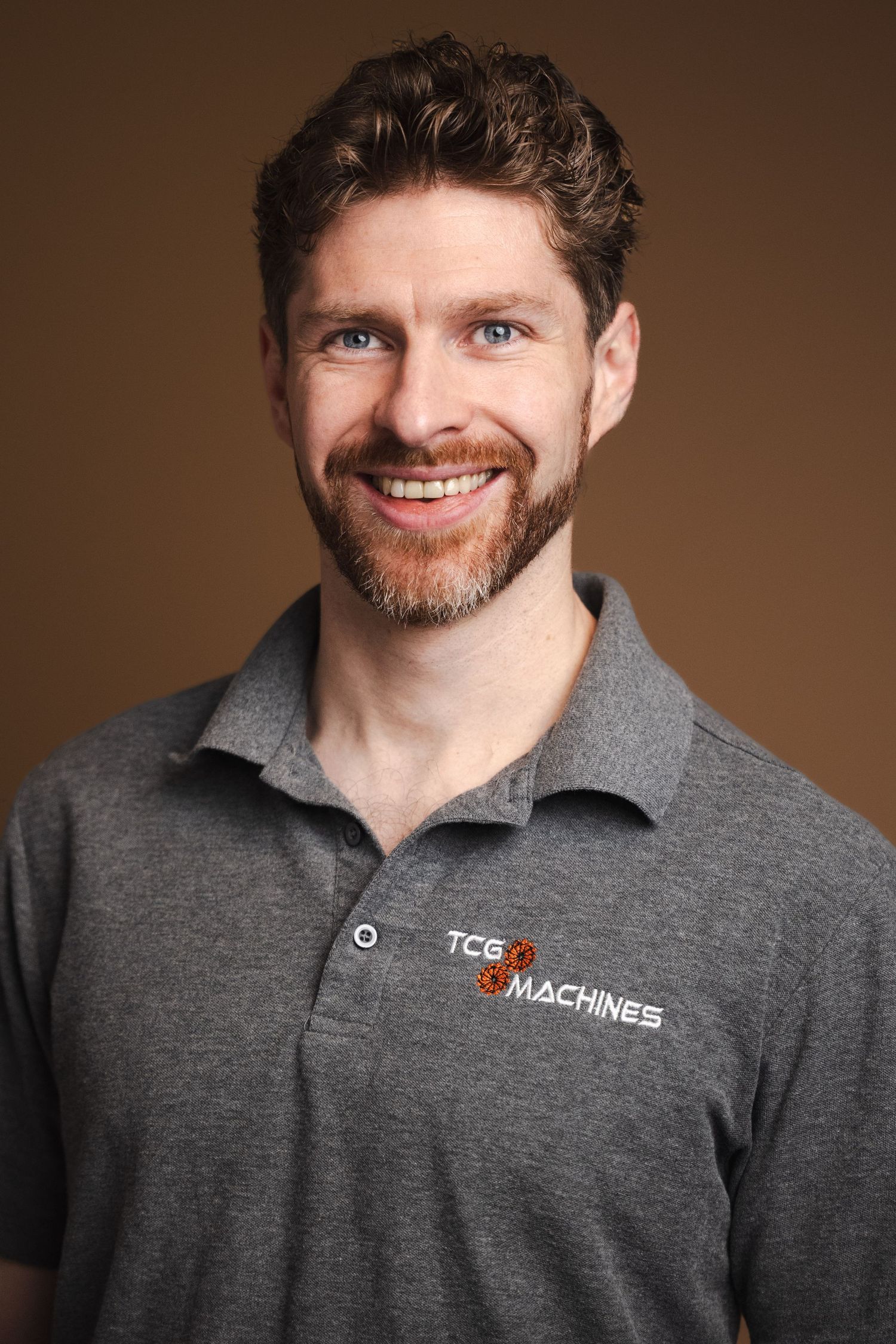 A person in a gray polo shirt with TCG Machines branding smiles warmly against a brown backdrop in a professional headshot series.