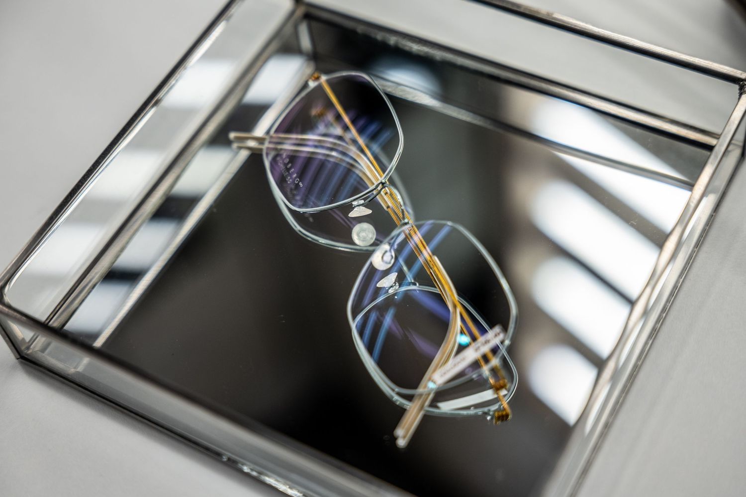 Elegant gold-framed eyeglasses with blue-tinted lenses displayed in a mirrored glass jewelry box.
