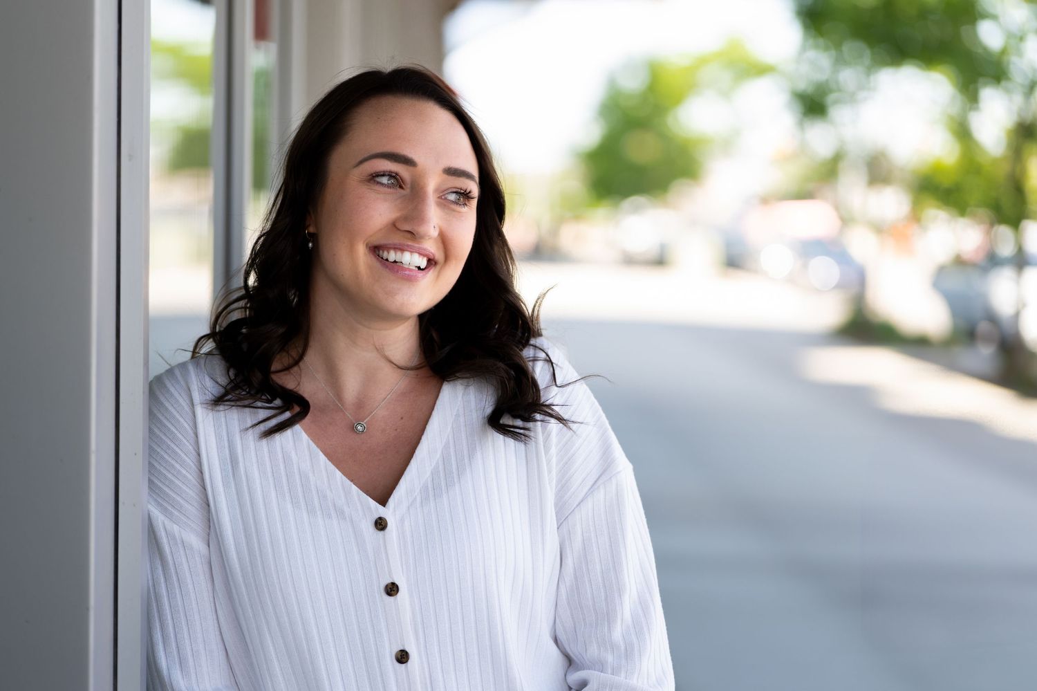 Professional woman in white button-up shirt smiles warmly while posing against a bright office background.