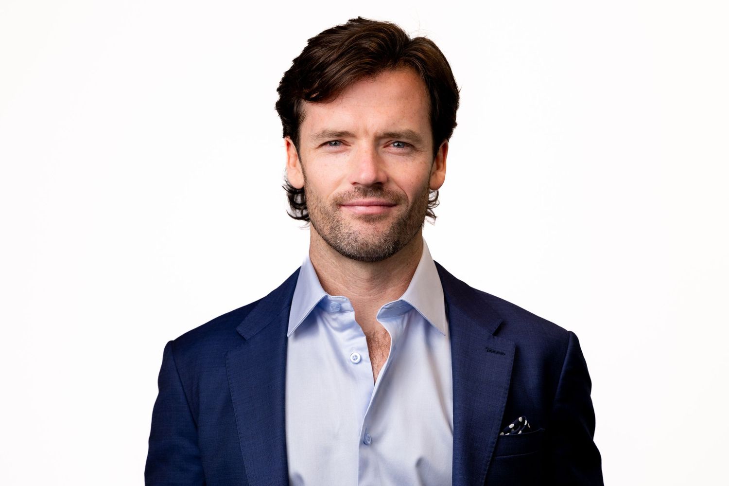 A person in a navy blue blazer and light blue button-down shirt smiles warmly against a white background.