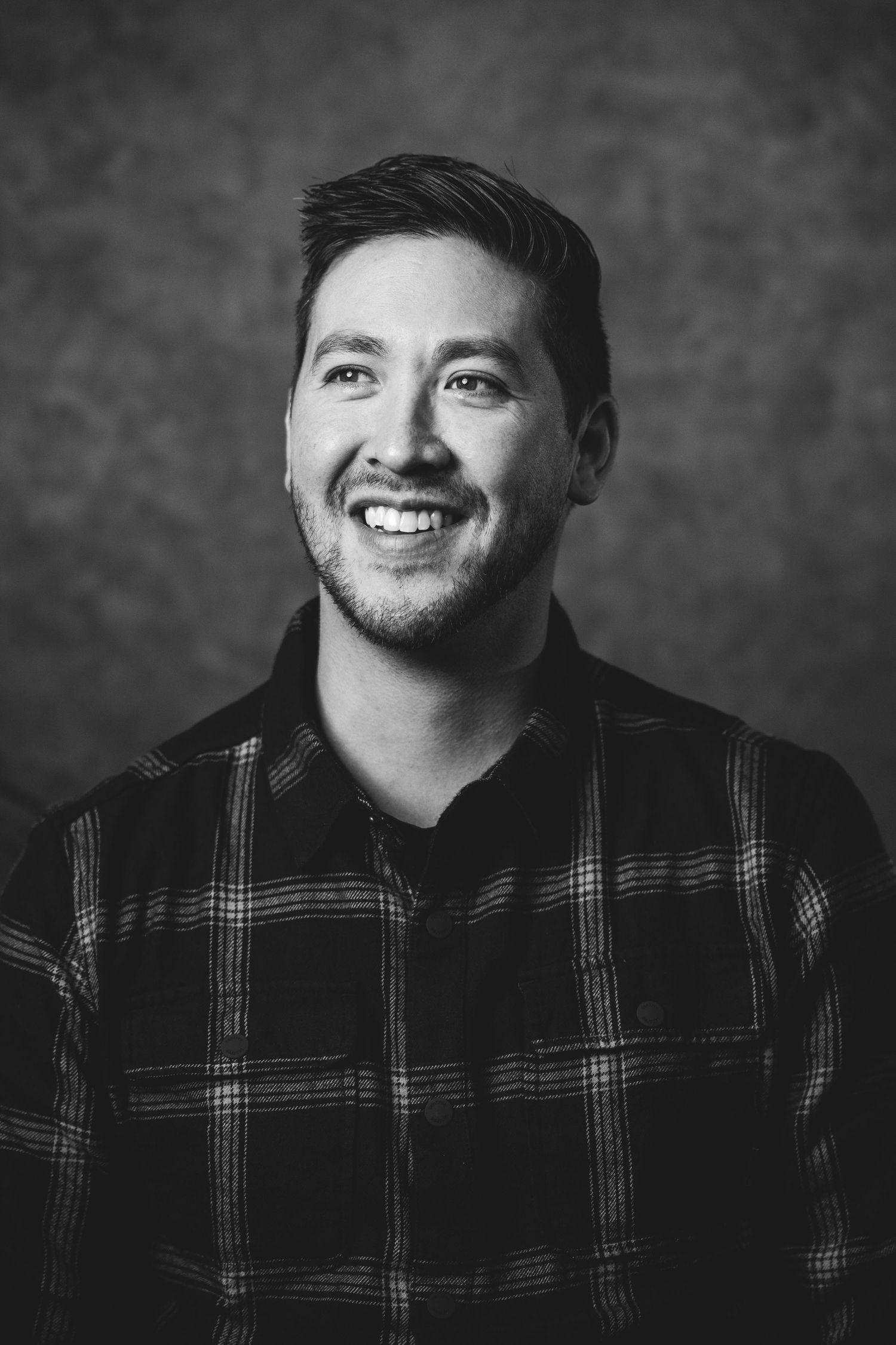 Smiling young man with stubble wearing a plaid shirt, black and white portrait photo.
