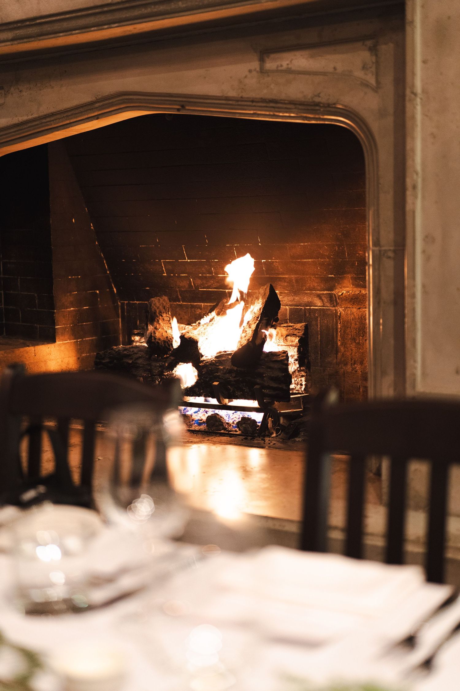 A cozy fireplace glows warmly behind an elegant dining table setting with white linens and dark wooden chairs.