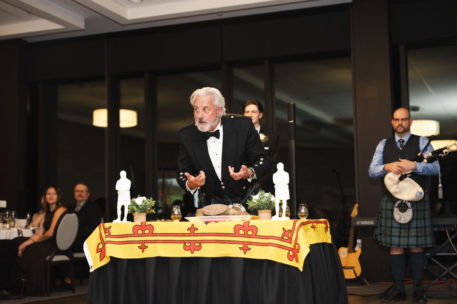 Elderly man in tuxedo speaks at decorated table during formal Scottish Burns Night dinner event with bagpiper in tartan.