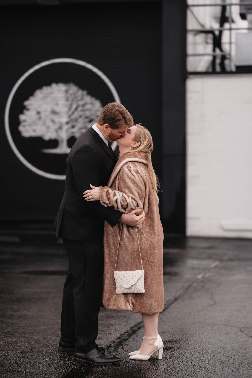 Two people embracing on a rainy day outside a building, one wearing a beige fur coat.