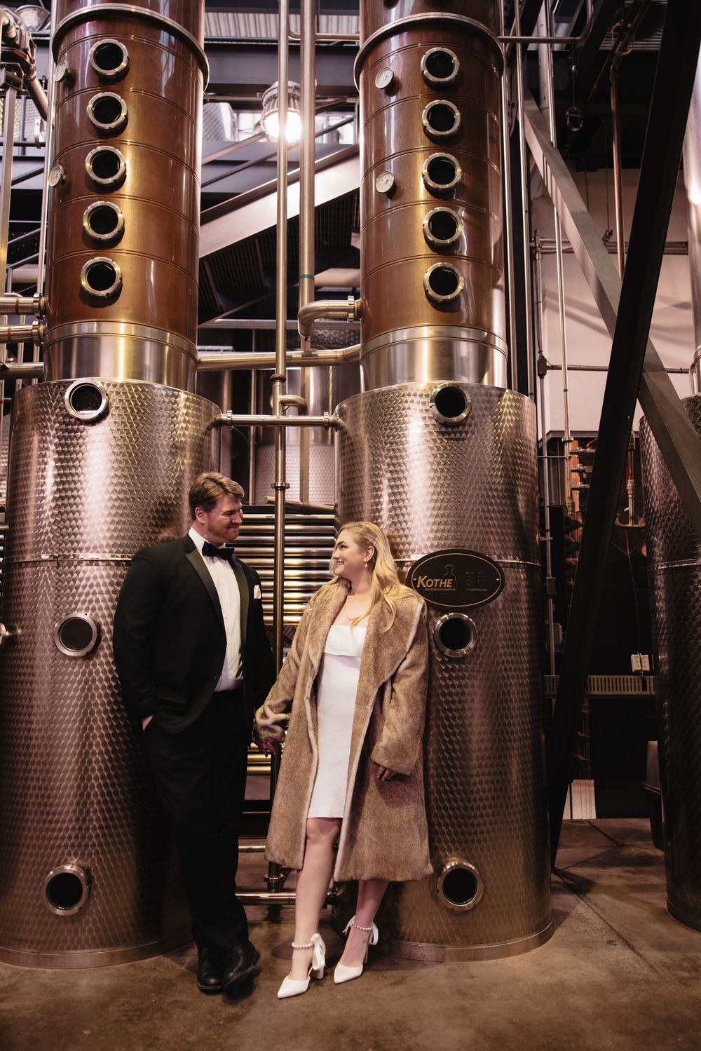 A couple stands between tall stainless steel distillery tanks in a modern production facility.