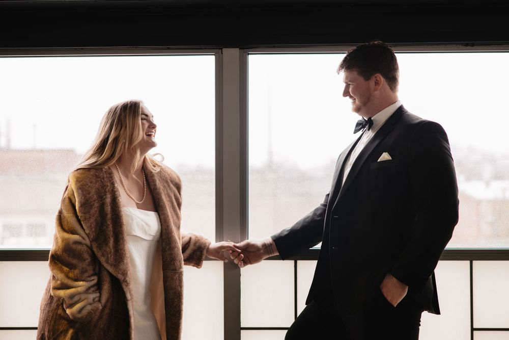 Silhouetted figures stand near large windows in an elegant indoor setting with natural light streaming in.