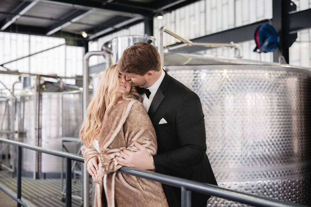 A couple in formal wedding attire share an intimate moment in an industrial space with stainless steel tanks.