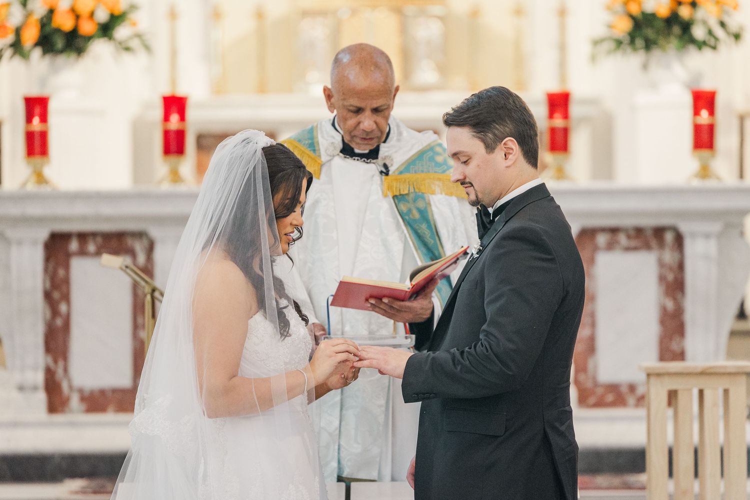 A wedding ceremony takes place in a church with white floral decorations and red banners hanging on the walls.