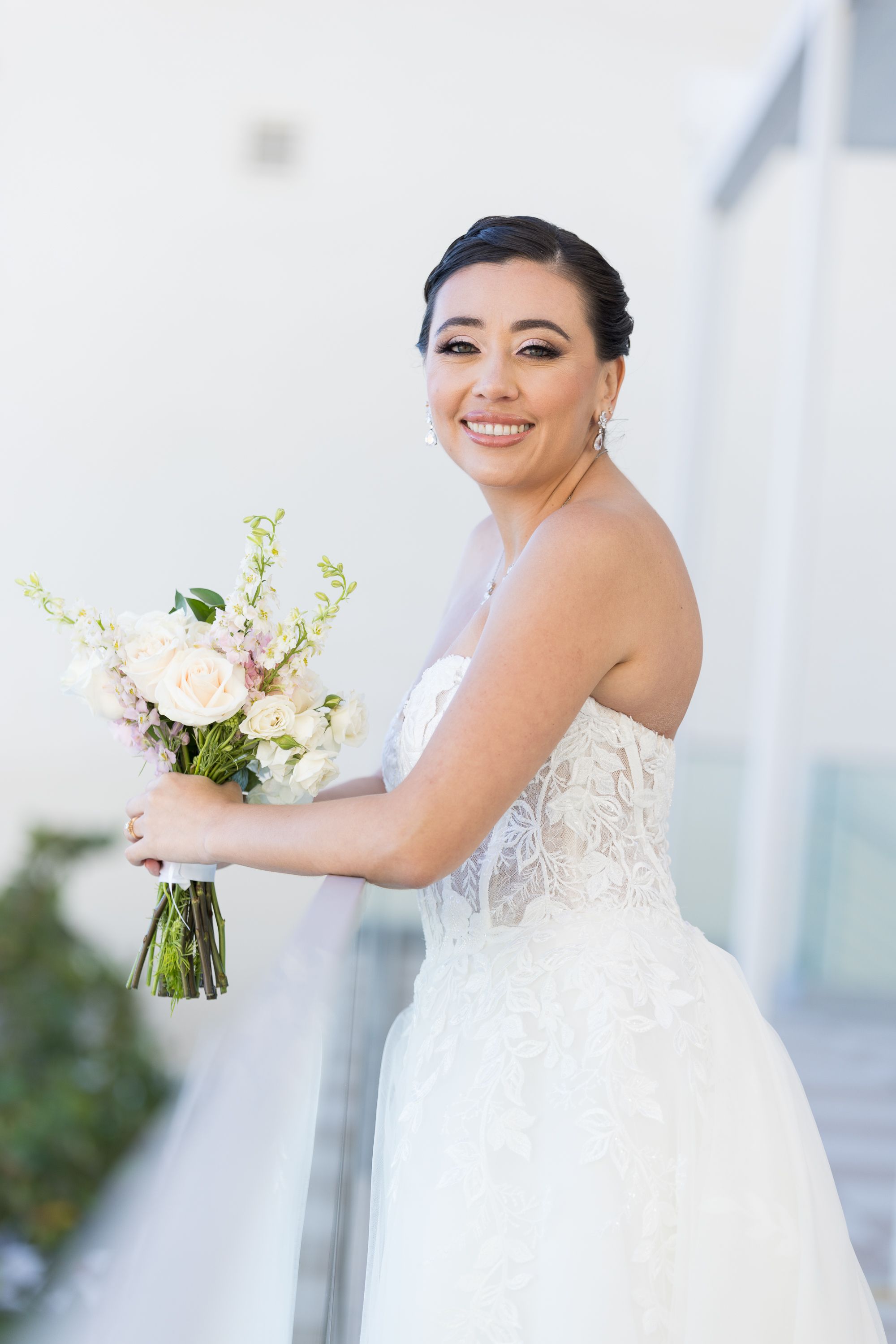 A bride in a strapless white lace wedding dress smiles while holding a bouquet of white flowers.