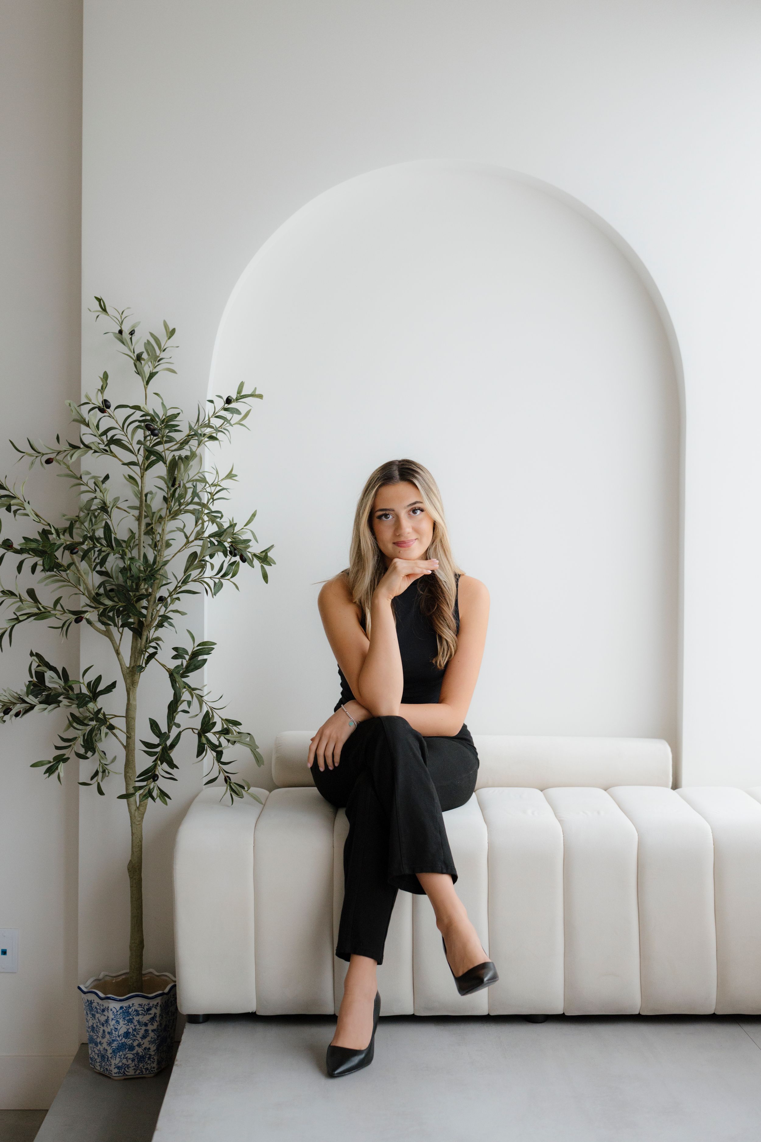 Woman in black outfit sitting elegantly on white bench near potted plant.