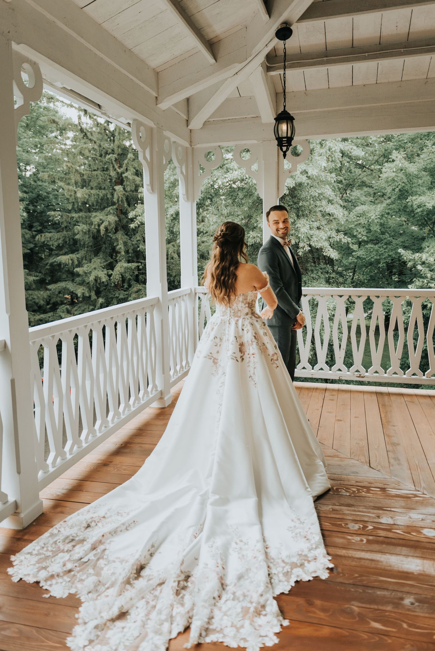 A bride and groom share a romantic moment on a covered porch with white railings and a long flowing lace wedding dress train.