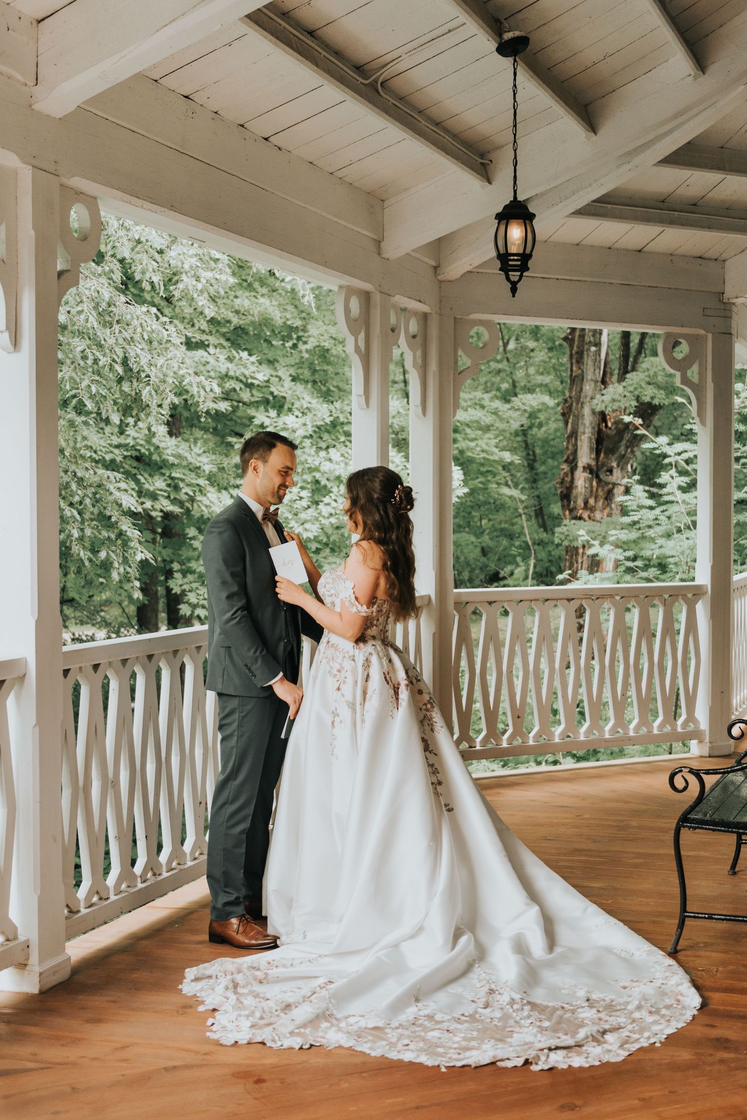 A couple shares a romantic moment on a rustic covered porch with white railings and lush greenery in the background.