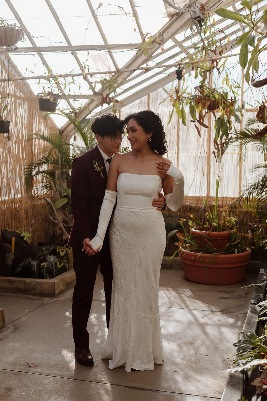 A couple shares an intimate dance moment in a sunlit greenhouse surrounded by lush potted plants and greenery.
