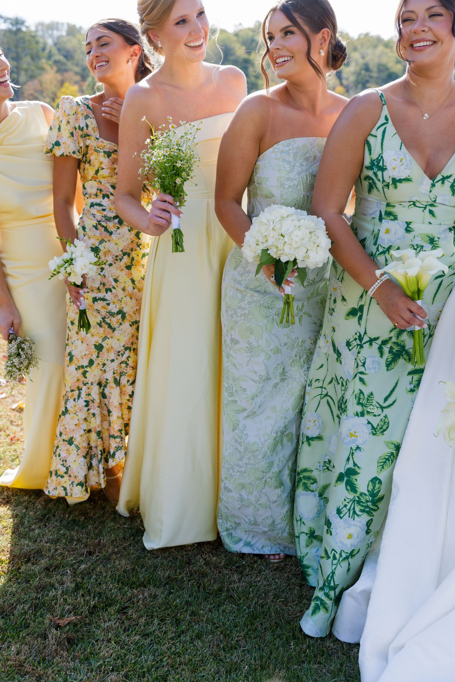 Bridal party in mix of yellow dresses and floral prints stands on sunny lawn with bride in flowing white gown with long train.