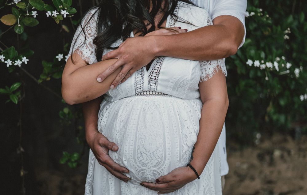 Close-up shot of hands cradling a pregnant belly in a white lace maternity dress against a natural backdrop.