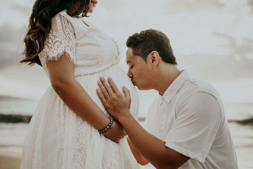 Intimate maternity moment captured on rocky beach with white lace dress.