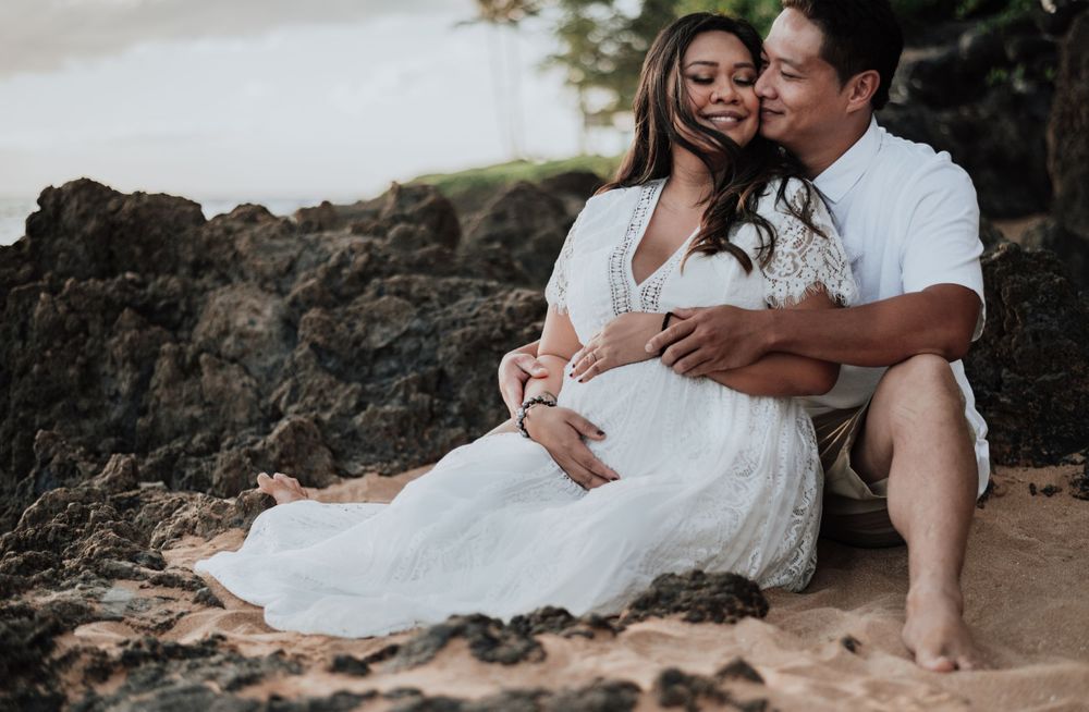 Romantic maternity photo of couple sitting on beach with rocky coastline at sunset.
