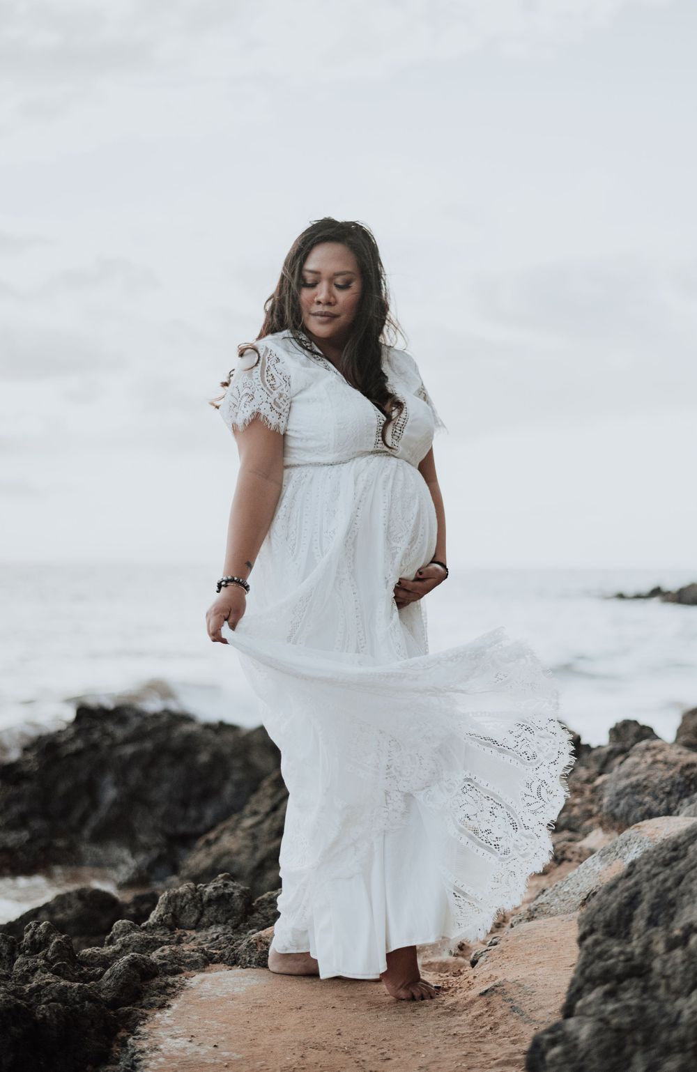 Ethereal maternity portrait featuring flowing white lace dress against dramatic coastal backdrop.
