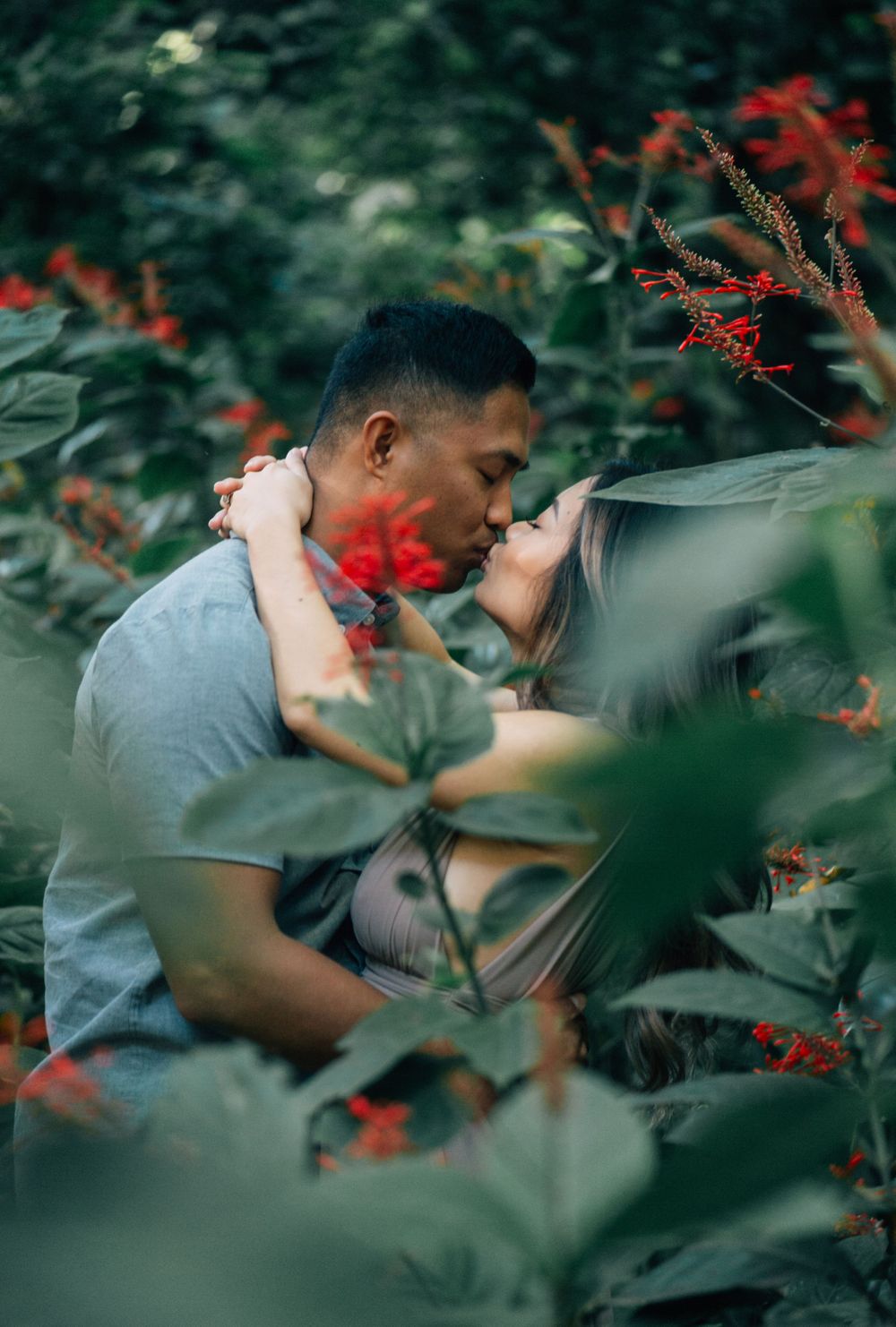 Two people share an intimate moment surrounded by red flowers and green leaves.